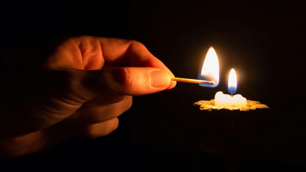 Una mano con un fiammifero accende una piccola candela, in una stanza buia. 